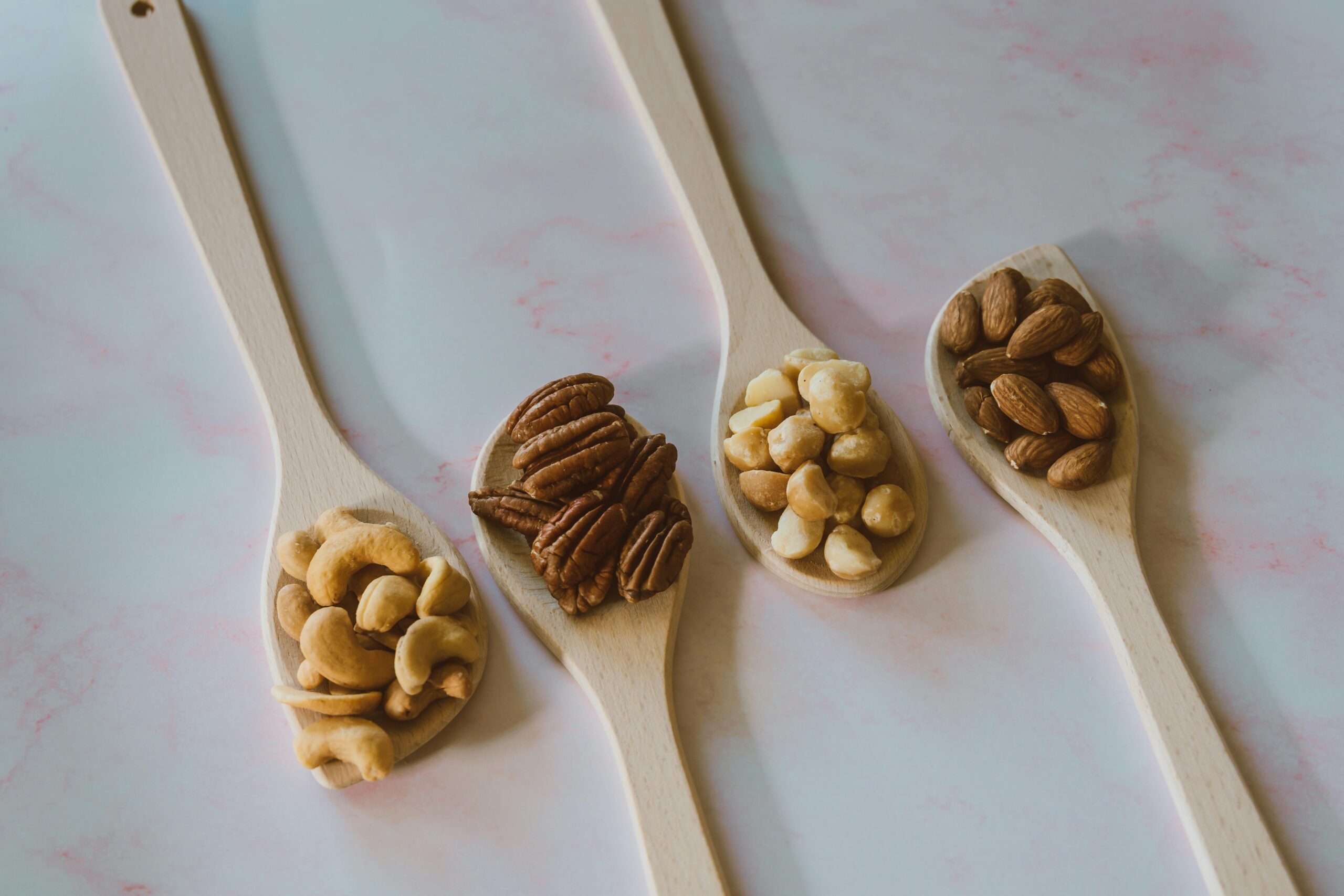 Close-up flat lay of various nuts like almonds and pecans in wooden spoons on a marble background.